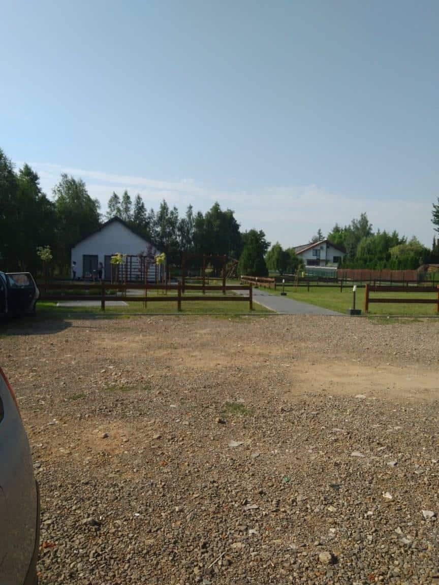 Żwirowy parking z drewnianym płotem, zielony trawnik i dwa białe domy otoczone drzewami pod czystym niebem.