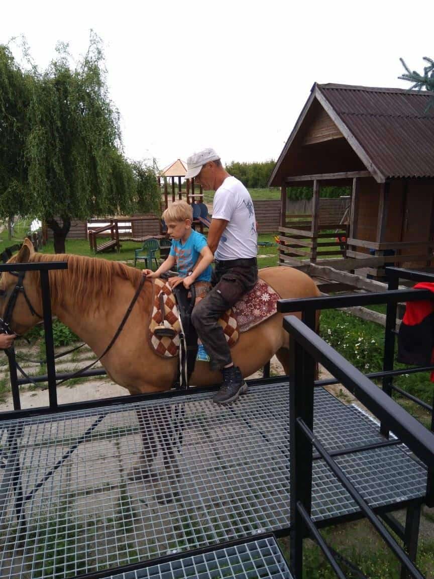 Dorosły pomaga młodemu chłopcu siedzącemu na osiodłanym koniu w pobliżu metalowej rampy i drewnianych budynków na zewnątrz.