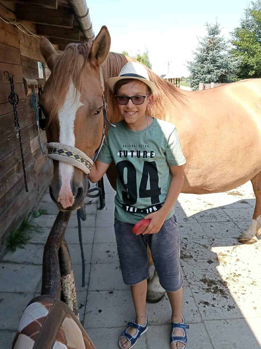 Chłopiec w okularach i kapeluszu stoi obok brązowego konia, uśmiechając się i trzymając czerwoną szczotkę, na zewnątrz w pobliżu drewnianego budynku.