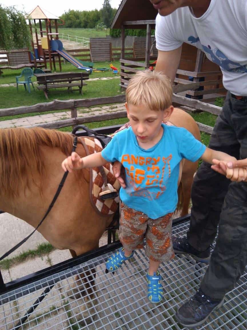 Młody chłopiec otrzymuje pomoc od osoby dorosłej, gdy stoi na metalowej platformie obok osiodłanego kucyka w parku na świeżym powietrzu z wyposażeniem placu zabaw w tle.