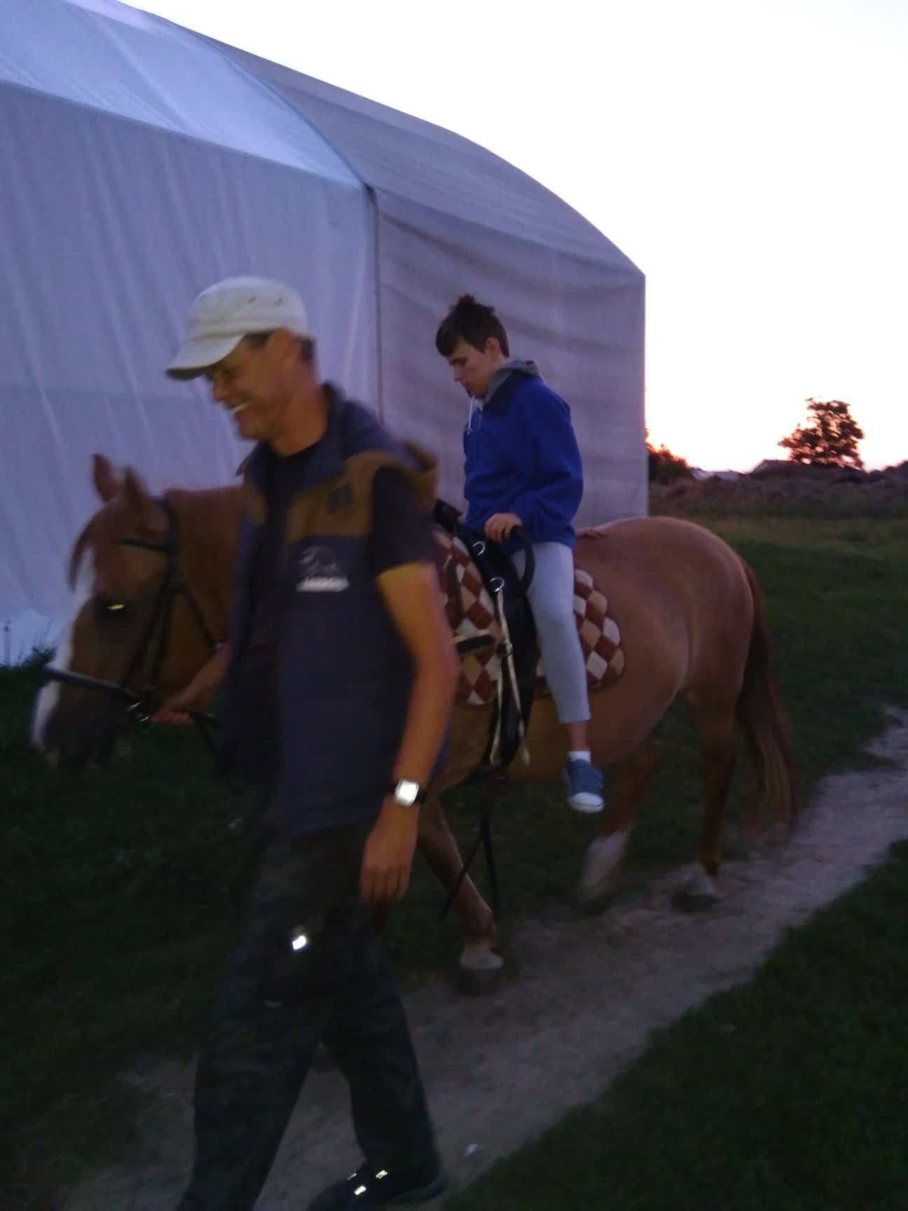 Mężczyzna prowadzi konia z dzieckiem w niebieskiej bluzie siedzącym na jego grzbiecie po polnej ścieżce w pobliżu białego namiotu o zmierzchu.