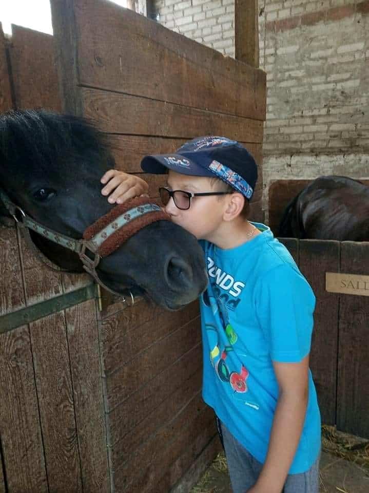 Chłopiec w niebieskiej koszuli i czapce całuje nos czarnego konia w drewnianej stajni.
