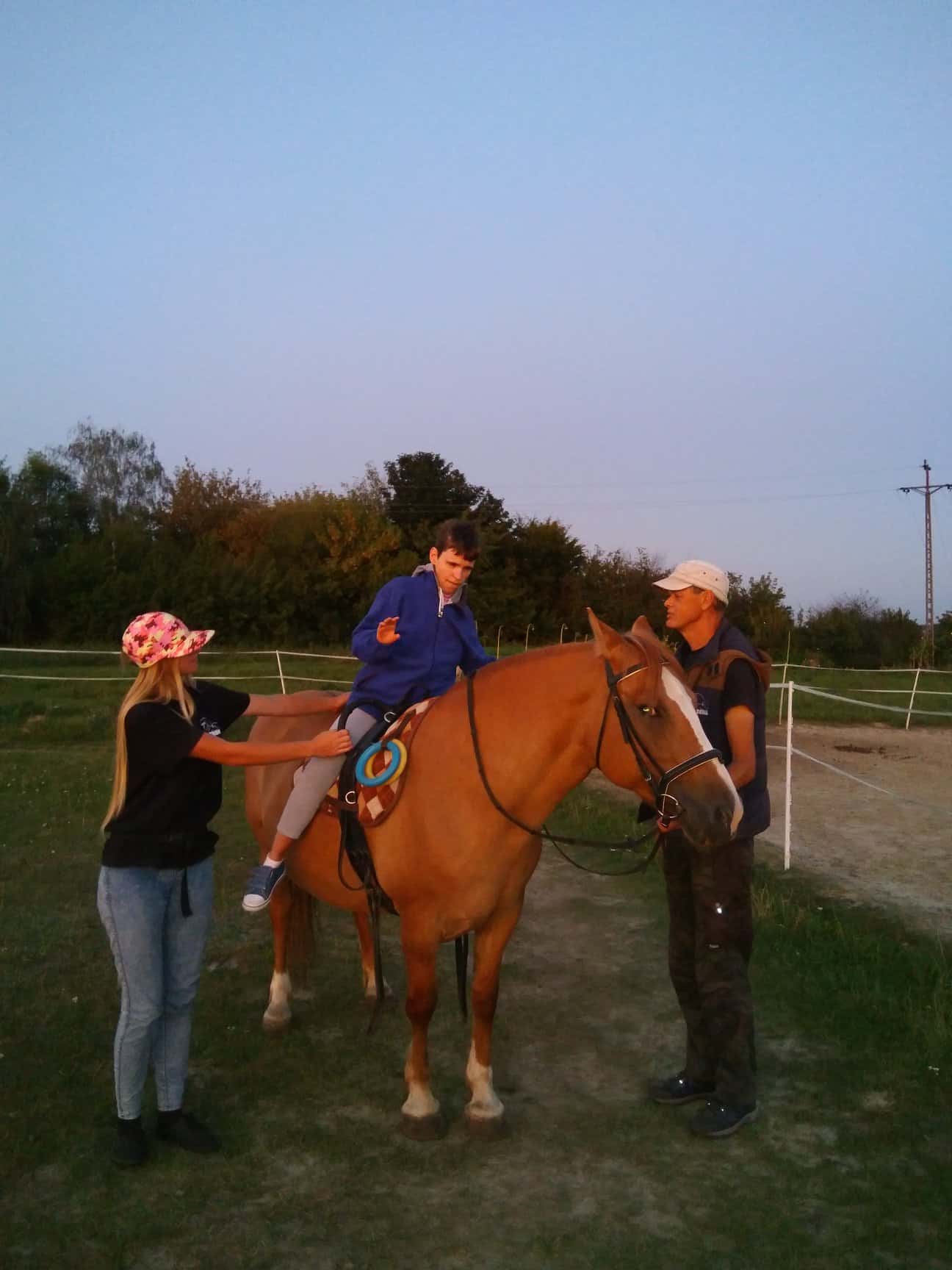 Młoda osoba siedzi na brązowym koniu, podczas gdy dwoje dorosłych stoi po obu stronach, pomagając i nadzorując w plenerze podczas zachodu słońca.