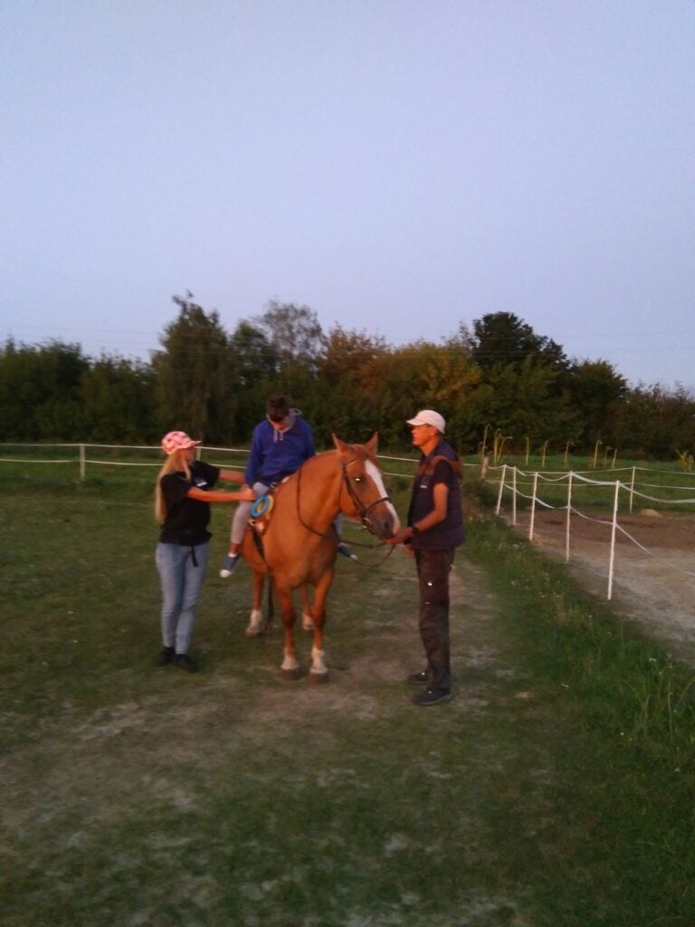 Osoba siedzi na brązowym koniu, podczas gdy inna osoba dostosowuje pozycję jeźdźca, a trzecia osoba trzyma lejce konia na zewnątrz.