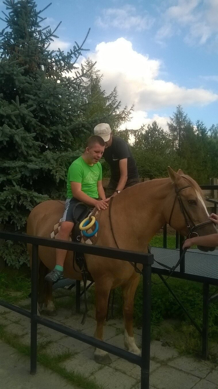 Dziecko w zielonej koszuli siedzi na jasnobrązowym koniu, w asyście dorosłego w czarnej koszuli i czapce, w plenerze z drzewami i zachmurzonym niebem.