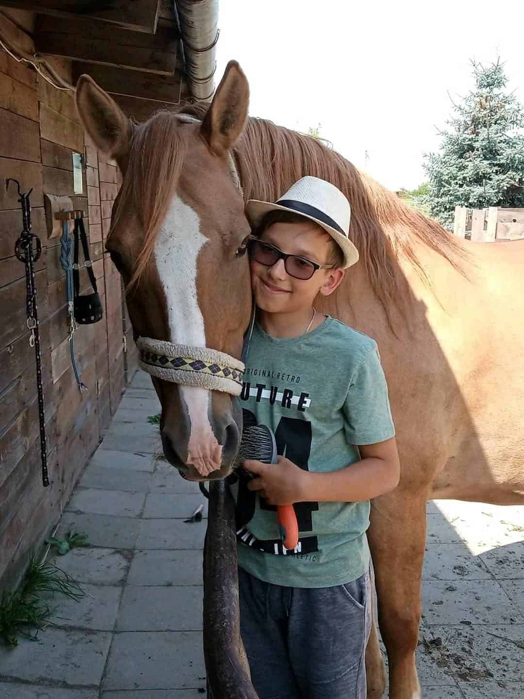 Chłopiec w okularach i kapeluszu stoi obok konia, uśmiecha się i trzyma szczotkę, a koń opiera głowę na jego ramieniu w stajni.