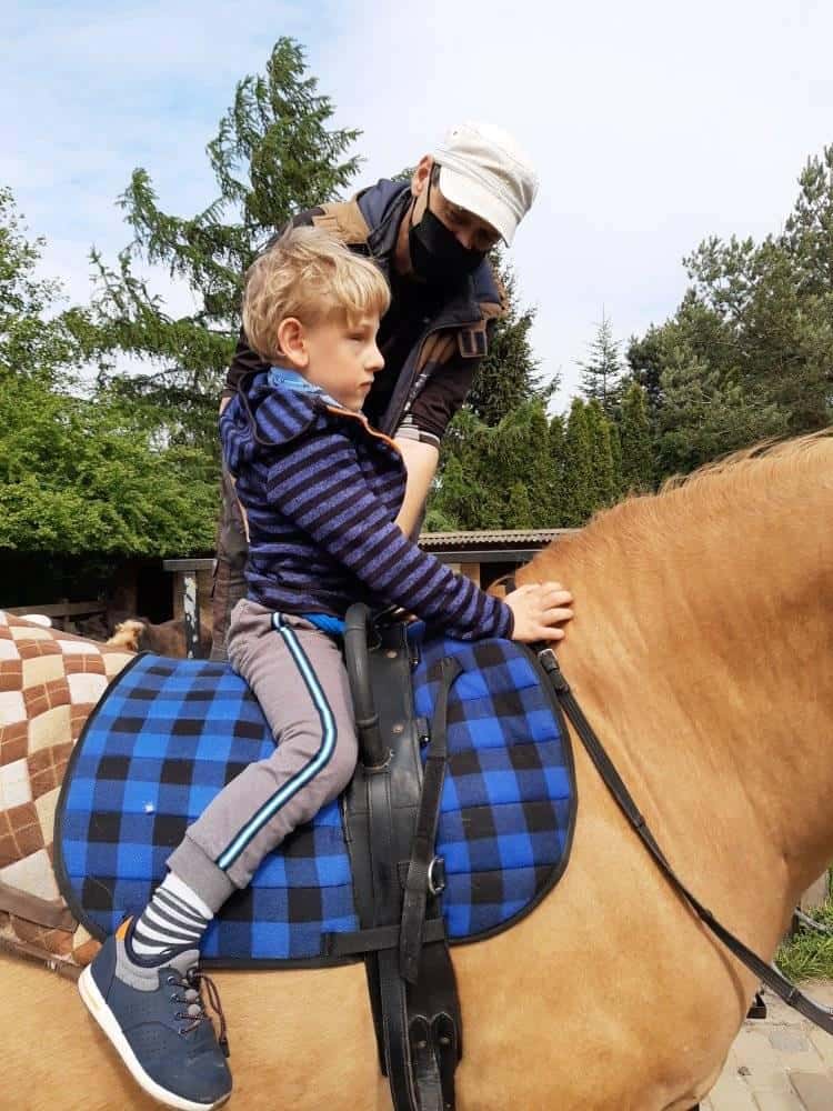Młody chłopiec siedzi na koniu z niebieskim siodłem w kratę, podczas gdy dorosły w masce i kapeluszu stoi obok niego, pomagając mu. W tle widoczne są drzewa.