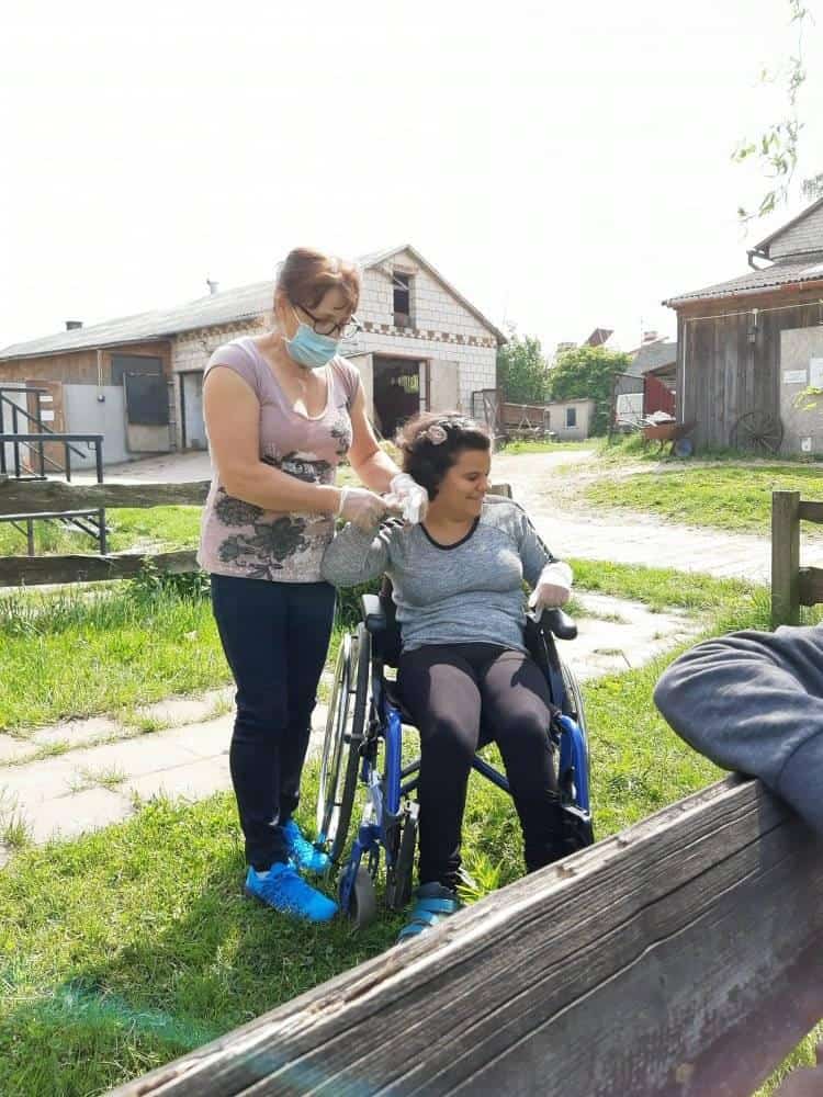 Kobieta w masce i rękawiczkach pomaga uśmiechniętej osobie siedzącej na wózku inwalidzkim na zewnątrz, na trawiastym terenie w pobliżu kilku budynków.