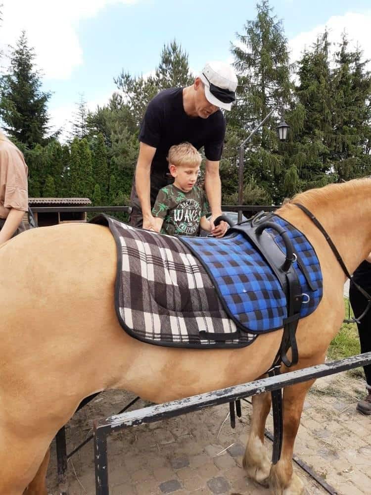 Dorosły pomaga młodemu chłopcu usiąść na osiodłanym jasnobrązowym koniu na zewnątrz, z drzewami i ogrodzeniem w tle.