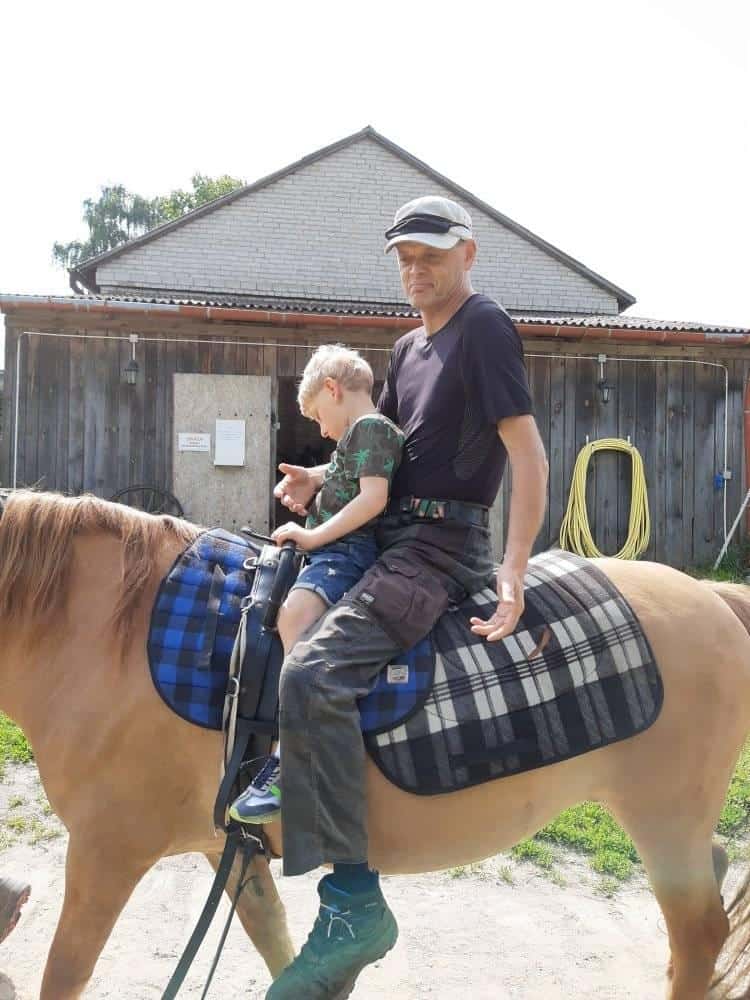 Dorosły i dziecko siedzą razem na brązowym koniu przed drewnianym budynkiem w słoneczny dzień.