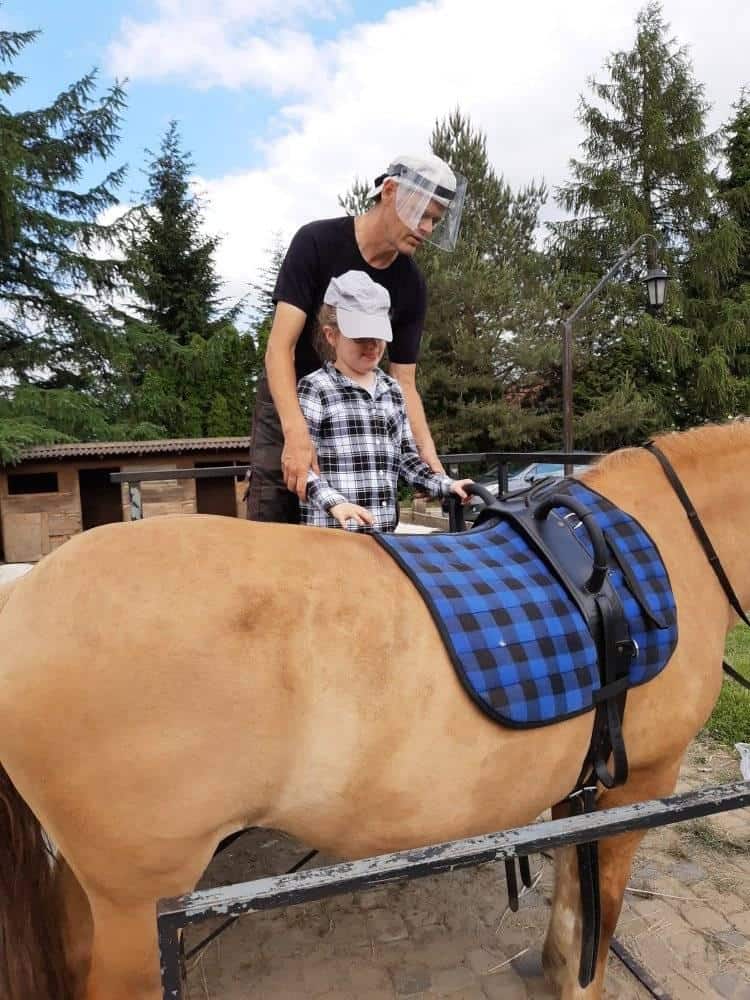 Dorosły pomaga dziecku siedzącemu na jasnobrązowym koniu z niebieskim siodłem w kratę. Oboje noszą czapki, a dorosły nosi również osłonę twarzy. W tle widać drzewa i stajnie.