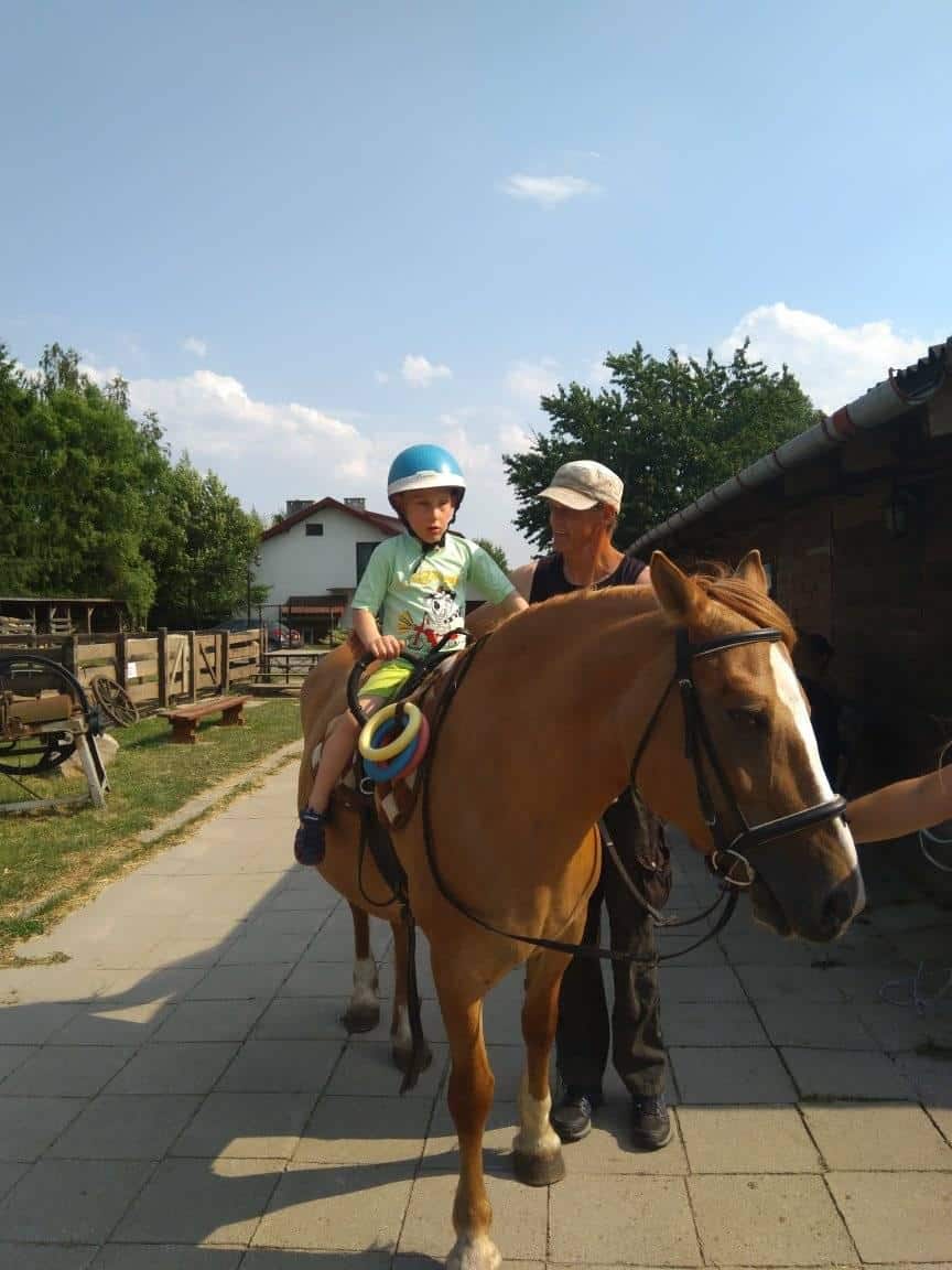Młody chłopiec w kasku siedzi na brązowym koniu, podczas gdy dorosły stoi obok niego, trzymając konia. Są na zewnątrz w pobliżu budynku i ogrodzenia w słoneczny dzień.