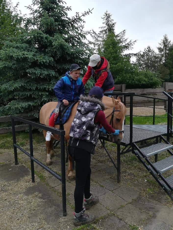 Osoba siedzi na małym brązowym koniu, podczas gdy dwoje dorosłych pomaga i prowadzi konia w pobliżu metalowej rampy na zewnątrz.