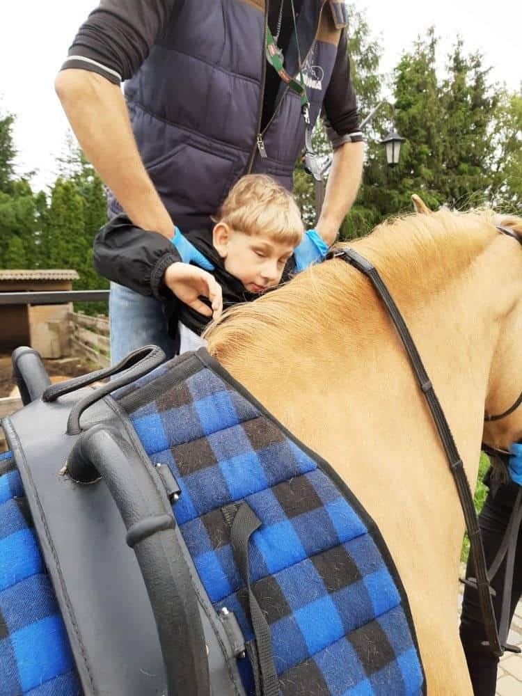 Małe dziecko w asyście osoby dorosłej siedzi na jasnobrązowym koniu z niebiesko-czarnym siodłem w kratkę. W tle widać drzewa i drewnianą konstrukcję.