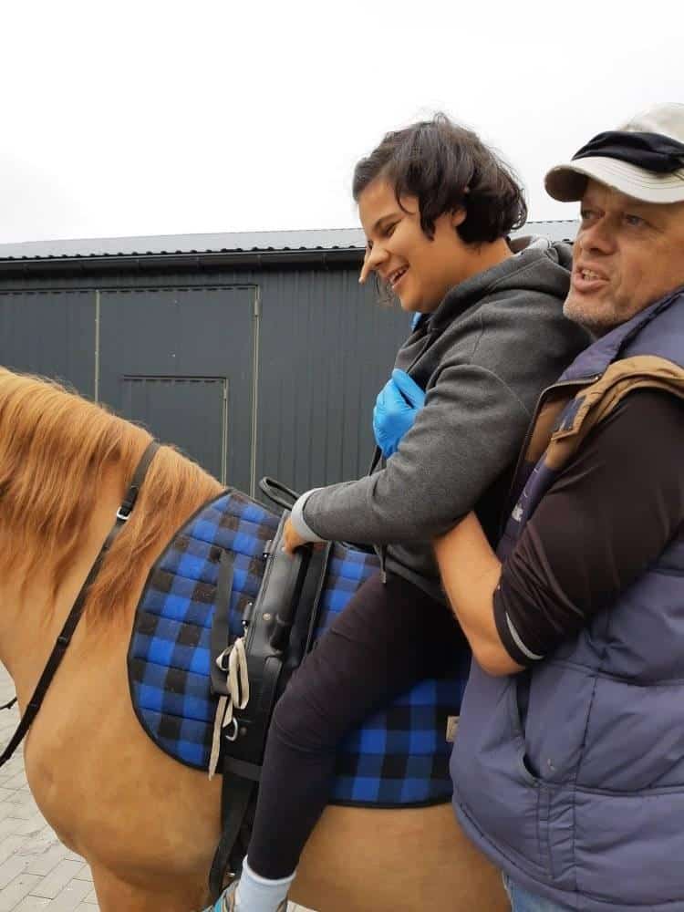 Osoba siedząca na koniu z niebieskim siodłem w kratę, w asyście innej osoby dorosłej stojącej obok. Oboje wydają się skupieni i zaangażowani. W tle widoczny jest budynek.