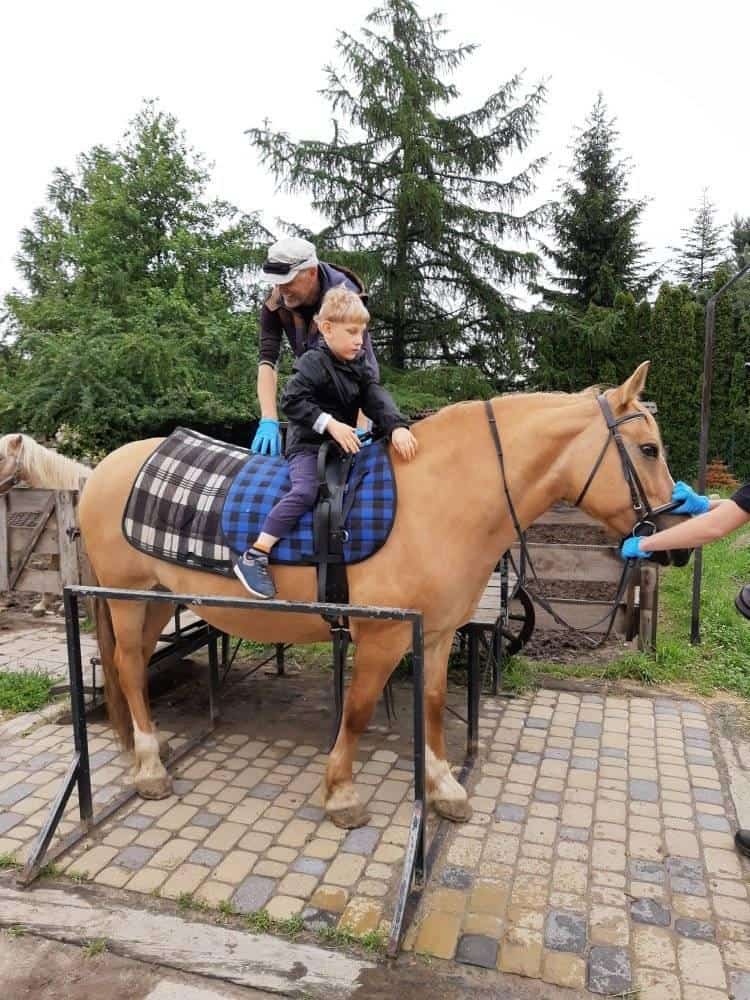 Mężczyzna pomaga młodemu chłopcu dosiąść osiodłanego konia, który stoi na ogrodzonym terenie na utwardzonej powierzchni na zewnątrz.