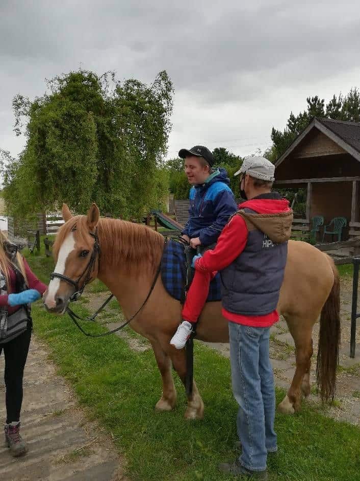 Osoba siedzi na jasnobrązowym koniu w siodle w kratę, w asyście dwóch dorosłych osób na zewnątrz w pobliżu drewnianego budynku w pochmurny dzień.