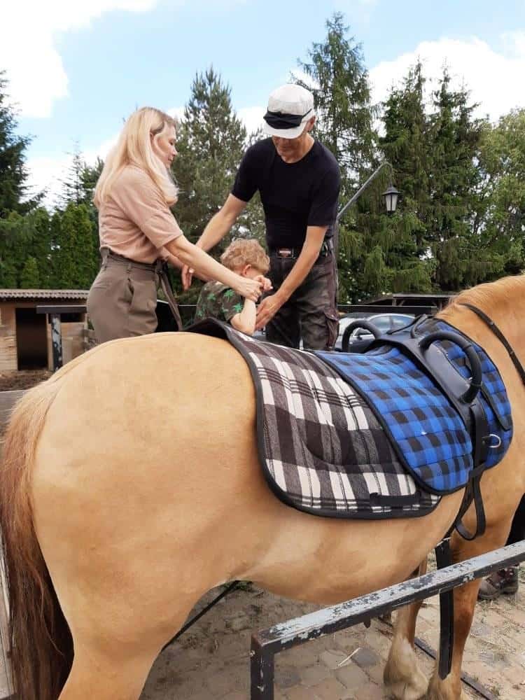 Dwoje dorosłych pomaga małemu dziecku usiąść na osiodłanym, opalonym koniu na zewnątrz. W tle widoczne są drzewa i ogrodzenie.