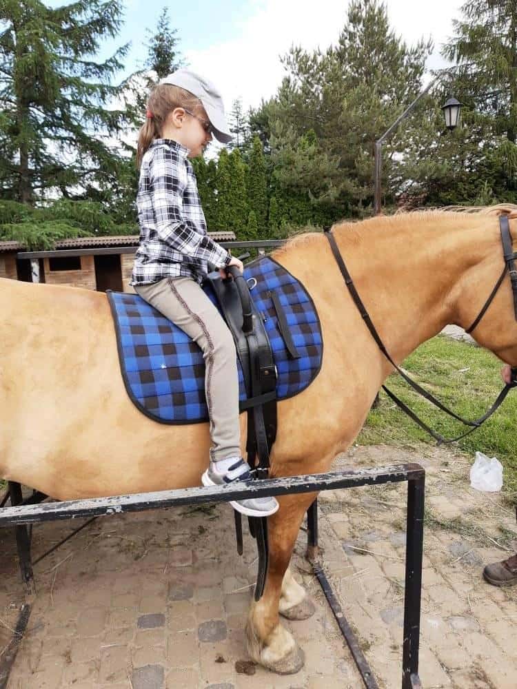 Młoda dziewczyna w czapce, koszuli w kratę i trampkach siedzi na osiodłanym koniu z niebieskim kocem w kratę w plenerze.