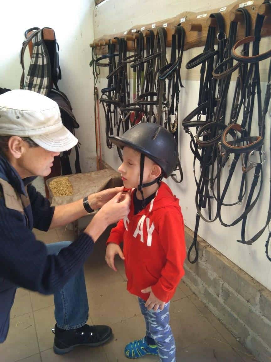 Dorosły zapina kask jeździecki małemu dziecku w stajni, za którymi na ścianie wiszą uzdy i sprzęt dla koni.