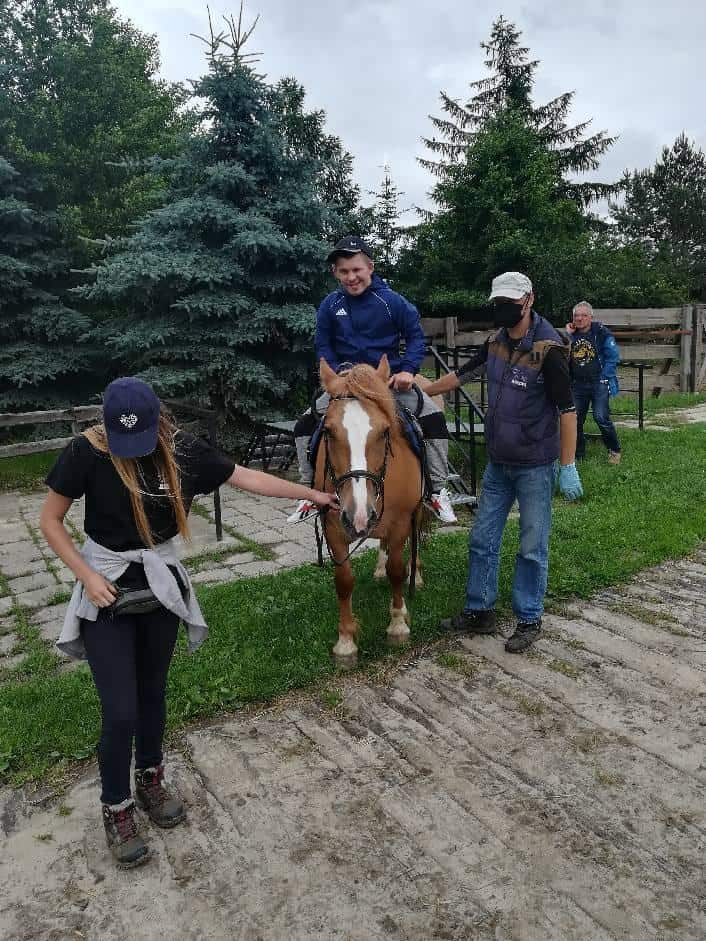 Mężczyzna siedzi na brązowym koniu, podczas gdy dwie osoby prowadzą konia po obu stronach; drzewa i ogrodzenie są w tle.