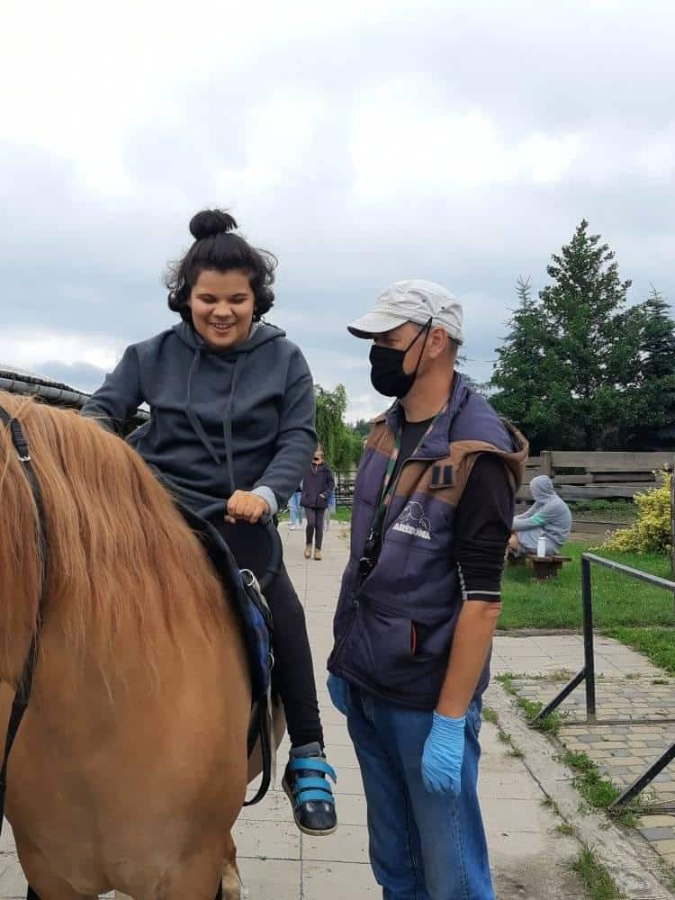 Młoda kobieta siedzi na koniu, uśmiechając się, podczas gdy mężczyzna w masce i rękawiczkach stoi obok niej na zewnątrz na brukowanej ścieżce.