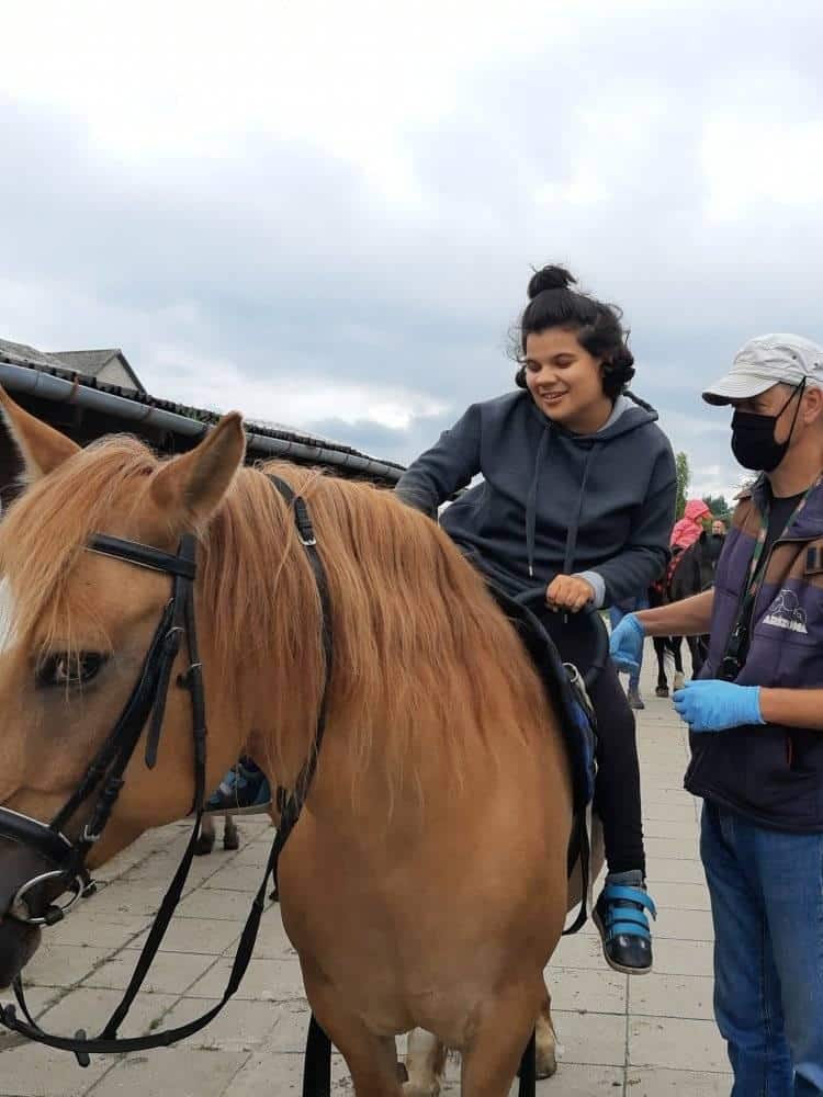 Młoda kobieta siedzi na jasnobrązowym koniu, podczas gdy mężczyzna w masce i rękawiczkach stoi obok nich na zewnątrz w pochmurny dzień.