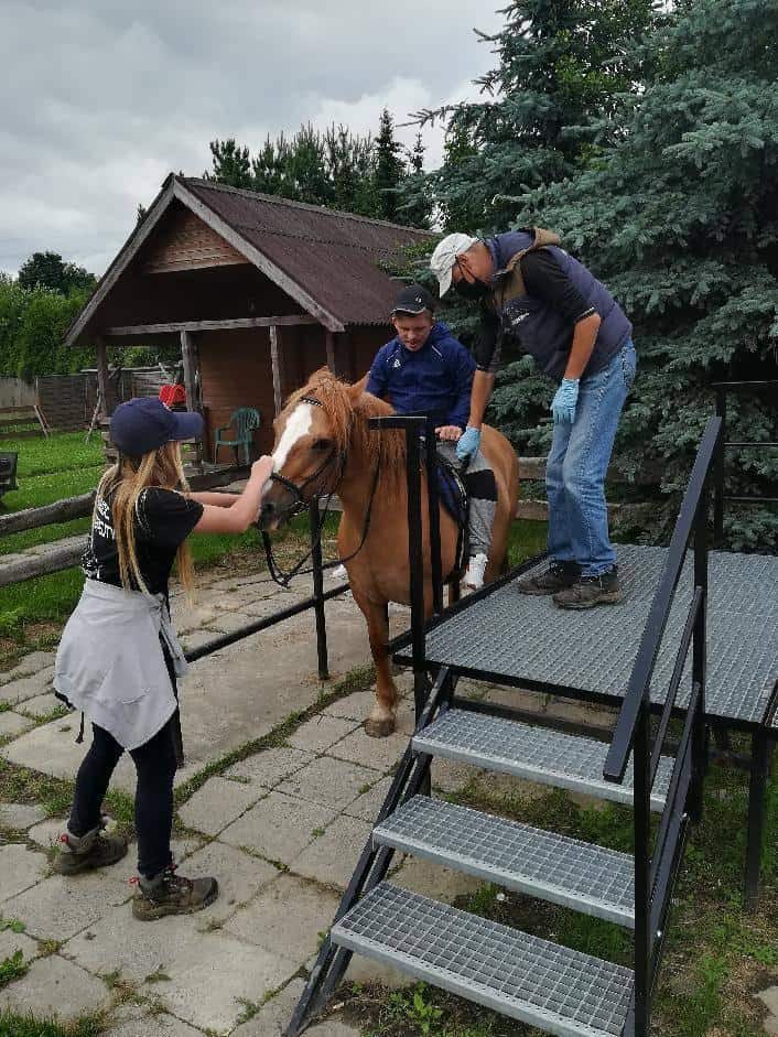 Trzy osoby pomagają mężczyźnie siedzącemu na brązowym koniu obok metalowej rampy na zewnątrz, w pobliżu drewnianej wiaty i drzew.