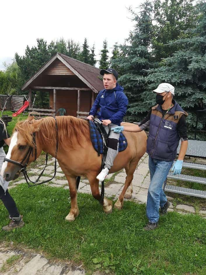 Młoda osoba z gipsem na prawej nodze jeździ na brązowym koniu na świeżym powietrzu w asyście dwóch dorosłych, z których jeden trzyma lejce, a drugi idzie obok.
