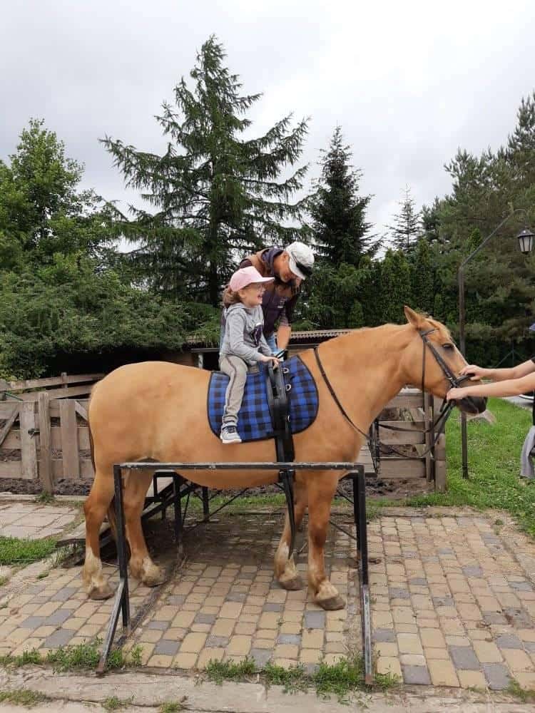 Dorosły pomaga małemu dziecku w różowej czapce usiąść na brązowym koniu z niebieską derką w kratę w plenerze.