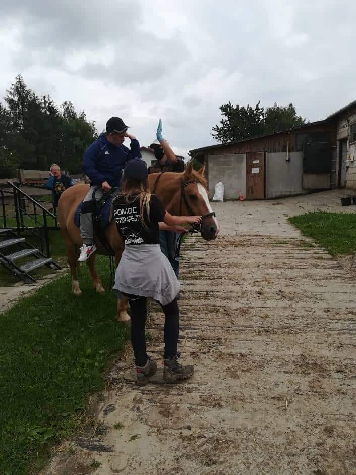 Osoba siedzi na brązowym koniu, podczas gdy dwie osoby pomagają, jedna trzyma lejce, a druga prowadzi z boku, na zewnątrz w pobliżu budynków gospodarczych.