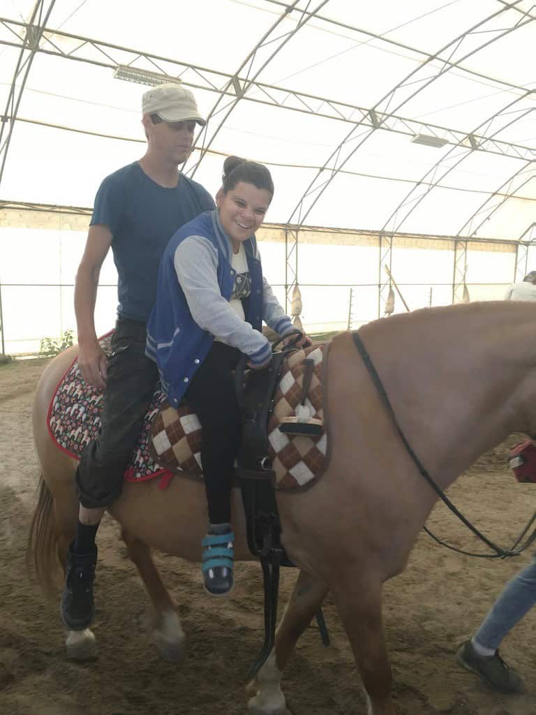 Mężczyzna i młoda kobieta jeżdżą razem na jasnobrązowym koniu na krytej arenie z brudną podłogą.