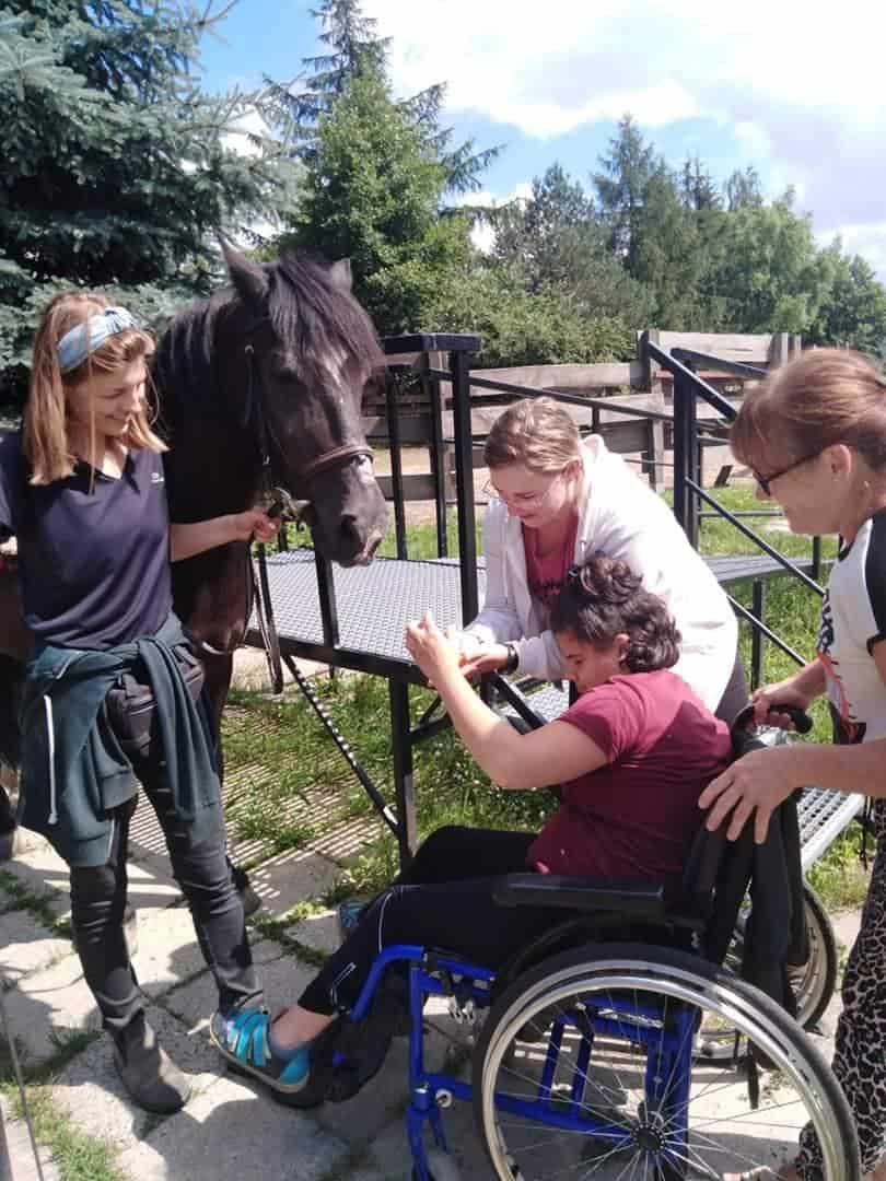 Kobieta na wózku inwalidzkim głaszcze czarnego konia z pomocą dwóch kobiet, podczas gdy inna kobieta trzyma wodze konia na zewnątrz.