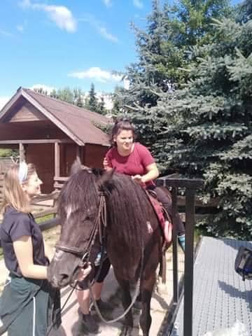 Kobieta siedzi na czarnym koniu, a obok niej stoi inna kobieta. W tle widać drzewa i drewniany budynek pod częściowo zachmurzonym niebem.