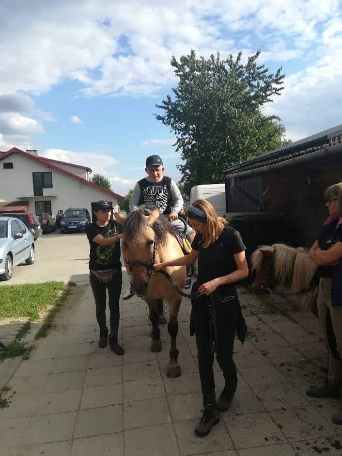 Osoba jedzie na brązowym koniu prowadzonym i wspomaganym przez trzy osoby po utwardzonej ścieżce w pobliżu budynków i zaparkowanych samochodów.
