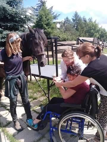 Trzy kobiety pomagają kobiecie na wózku inwalidzkim podczas interakcji z koniem na świeżym powietrzu w słoneczny dzień.