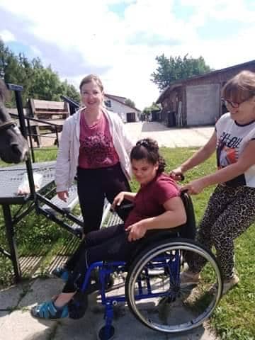 Uśmiechnięta dziewczyna na wózku inwalidzkim jest wspomagana przez dwie kobiety na zewnątrz w pobliżu rampy, z koniem widocznym po lewej stronie obrazu.