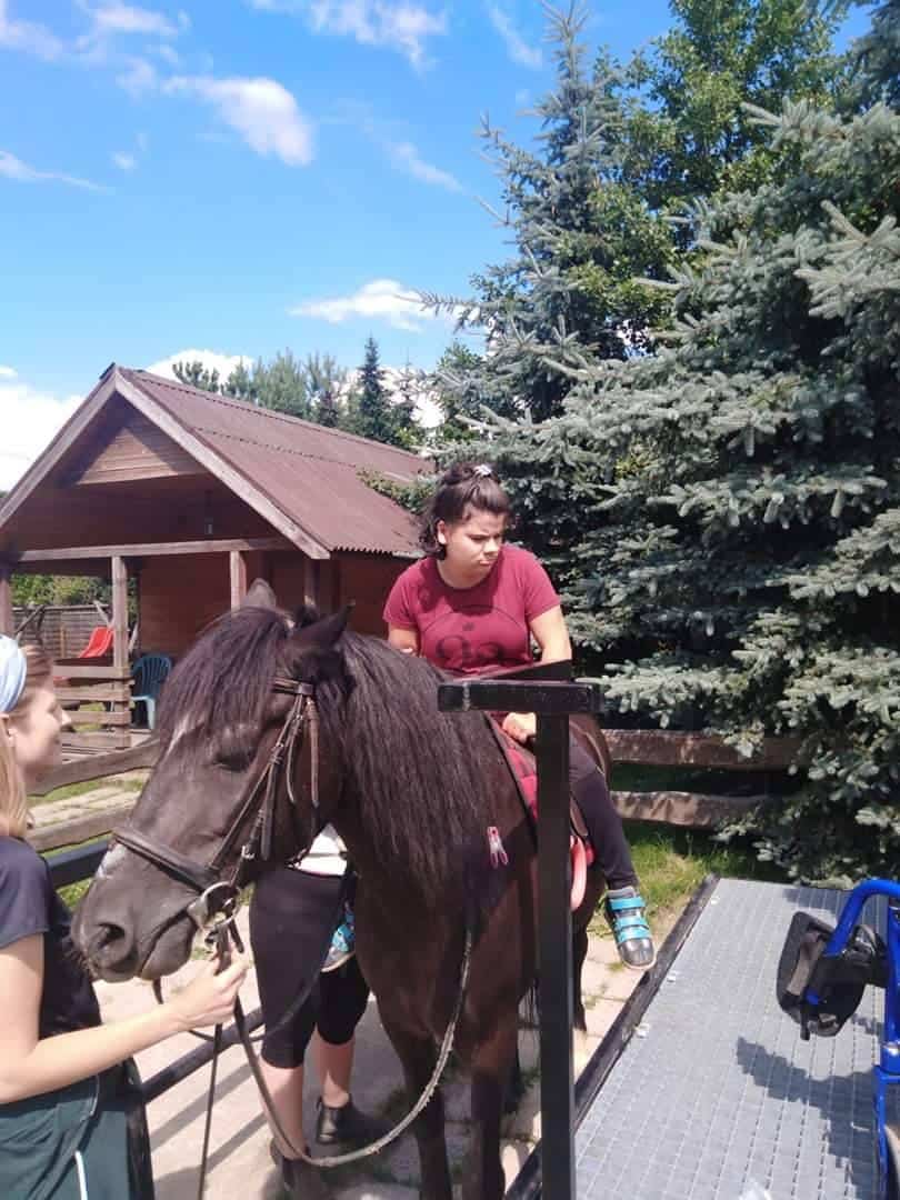 Osoba siedzi na ciemnym koniu w pobliżu drewnianego budynku, z dwiema osobami asystującymi, pod słonecznym niebem z sosnami w tle.