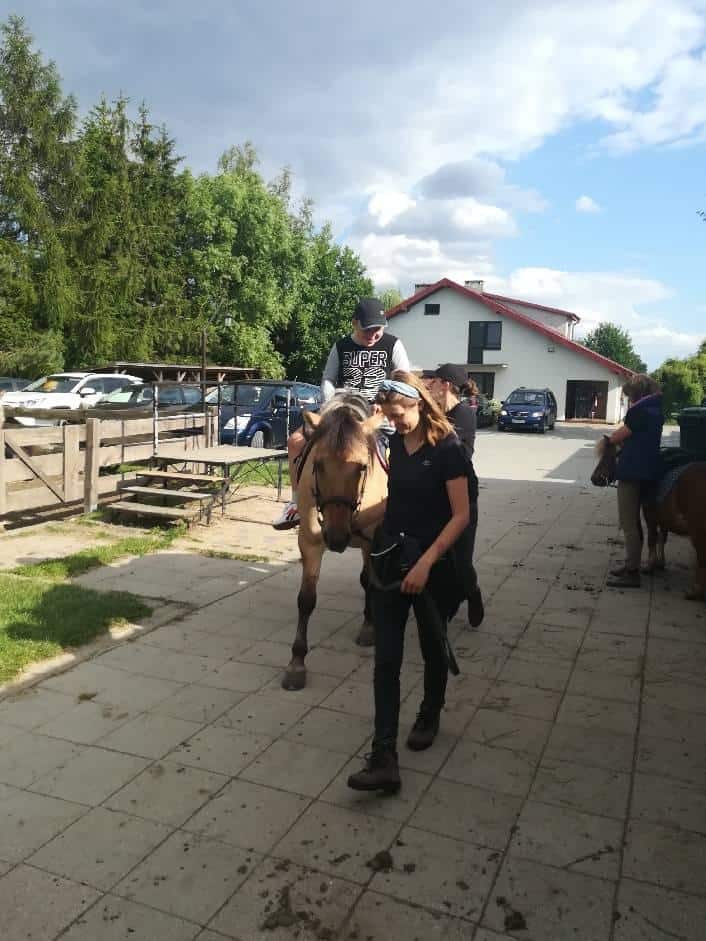 Osoba w kasku jedzie na koniu prowadzonym przez inną osobę po utwardzonej ścieżce w pobliżu domu z zaparkowanymi samochodami w tle.