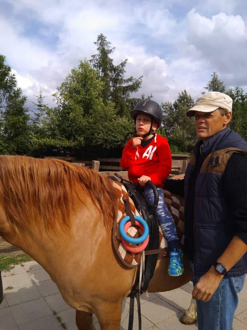 Dziecko w kasku i czerwonej bluzie siedzi na brązowym koniu w towarzystwie dorosłego mężczyzny na zewnątrz w pobliżu drzew pod zachmurzonym niebem.