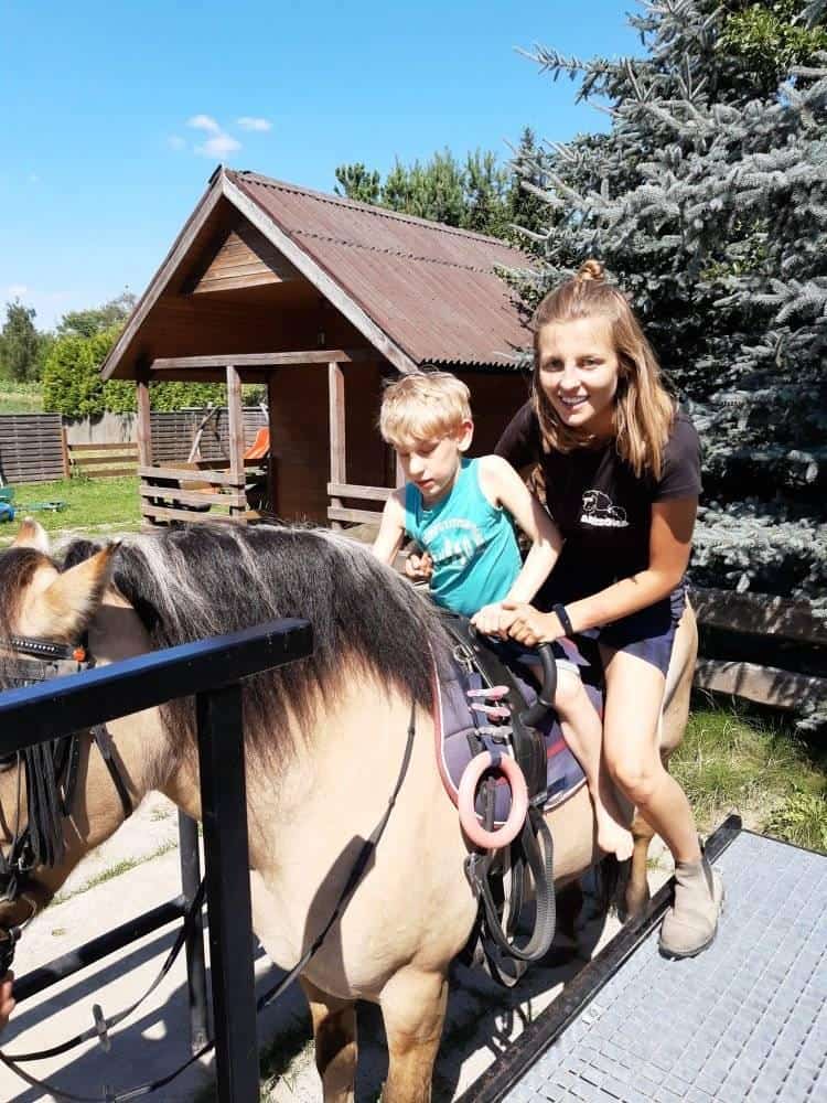 Kobieta pomaga młodemu chłopcu jadącemu na małym koniu na zewnątrz w pobliżu drewnianej szopy i drzew w słoneczny dzień.
