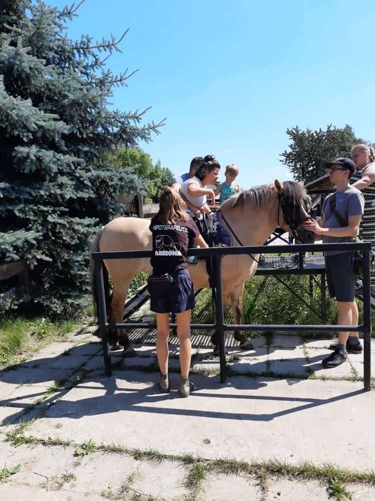 Dziecko siedzi na koniu w asyście dorosłych i członka personelu w czarnej koszuli z napisem "Hipoterapia". Są na zewnątrz w pobliżu drzew i ogrodzenia w słoneczny dzień.