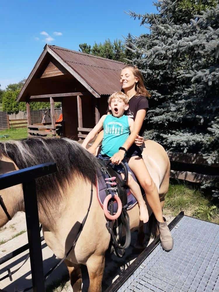 Młoda kobieta i młody chłopiec siedzą razem na koniu w pobliżu drewnianej szopy i sosny. Chłopiec wygląda na zaskoczonego, a kobieta się uśmiecha.