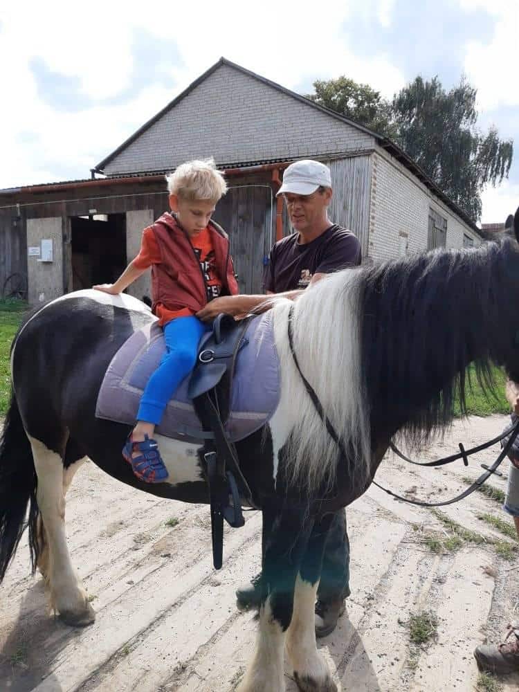 Dorosły pomaga młodemu chłopcu siedzącemu na czarno-białym koniu w pobliżu stodoły w częściowo pochmurny dzień.