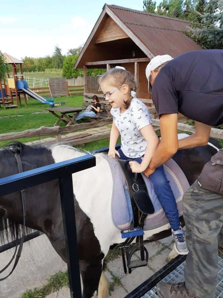 Młoda dziewczyna siedzi na małym koniu, a dorosły jej pomaga. W tle znajduje się drewniany dom, stół piknikowy i wyposażenie placu zabaw.
