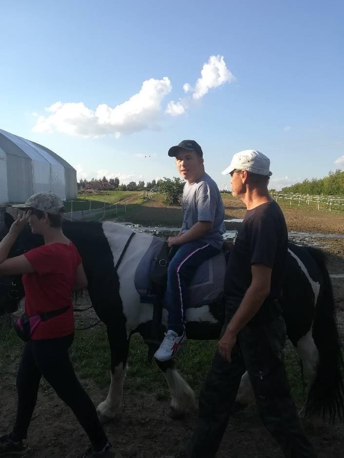 Chłopiec jedzie na czarno-białym koniu w towarzystwie dwóch dorosłych idących obok niego po polnej ścieżce w pobliżu szklarni.