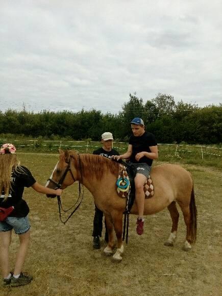 Osoba siedzi na brązowym koniu, podczas gdy dwie inne osoby stoją w pobliżu, trzymając lejce na trawiastym terenie.