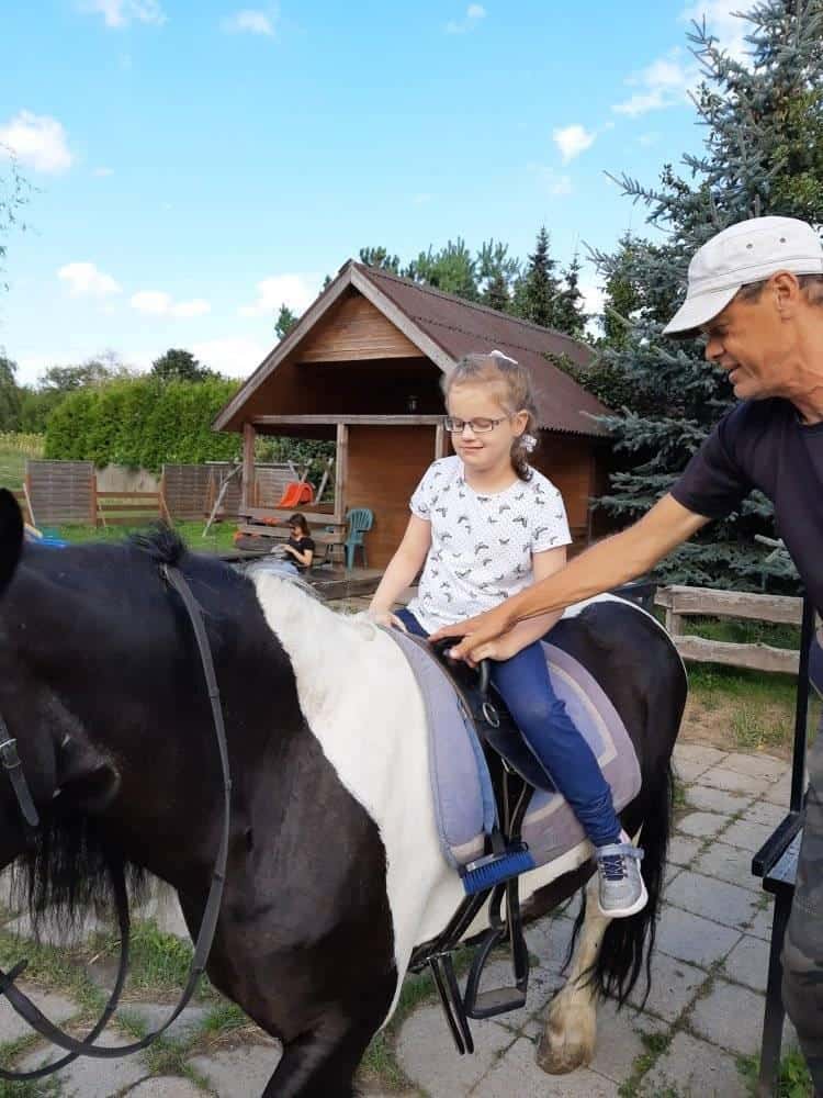 Mężczyzna pomaga młodej dziewczynie w okularach, która siedzi na czarno-białym koniu na podwórku z drewnianą szopą w tle.