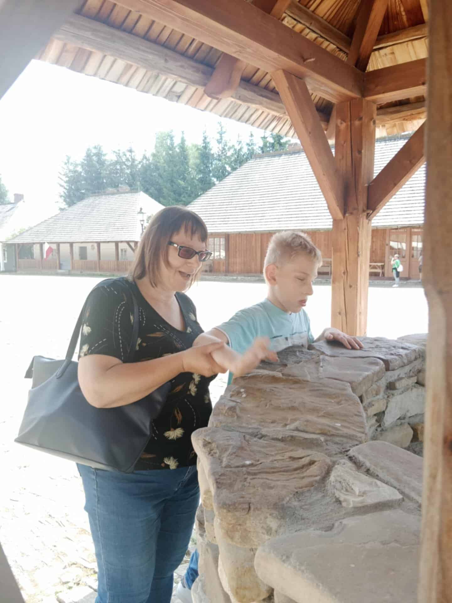 Kobieta i chłopiec stoją przy kamiennej studni pod drewnianą wiatą, dotykając kamieni. W tle widoczne są drewniane budynki i drzewa w słoneczny dzień.