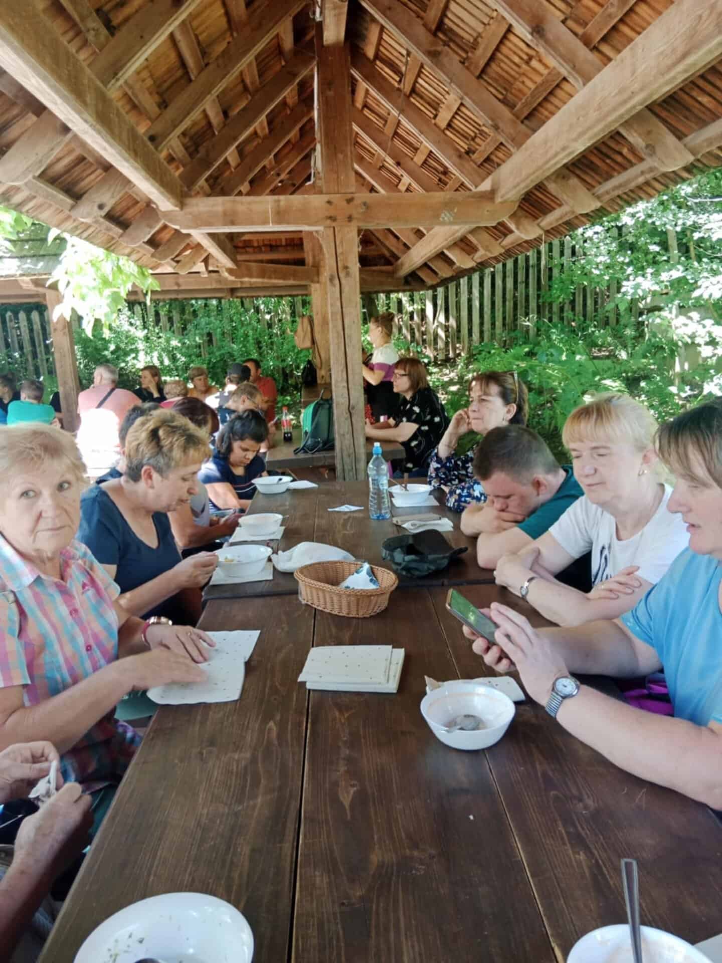 Grupa ludzi siedzi przy długim drewnianym stole pod drewnianą wiatą, jedząc i patrząc na menu lub telefony.